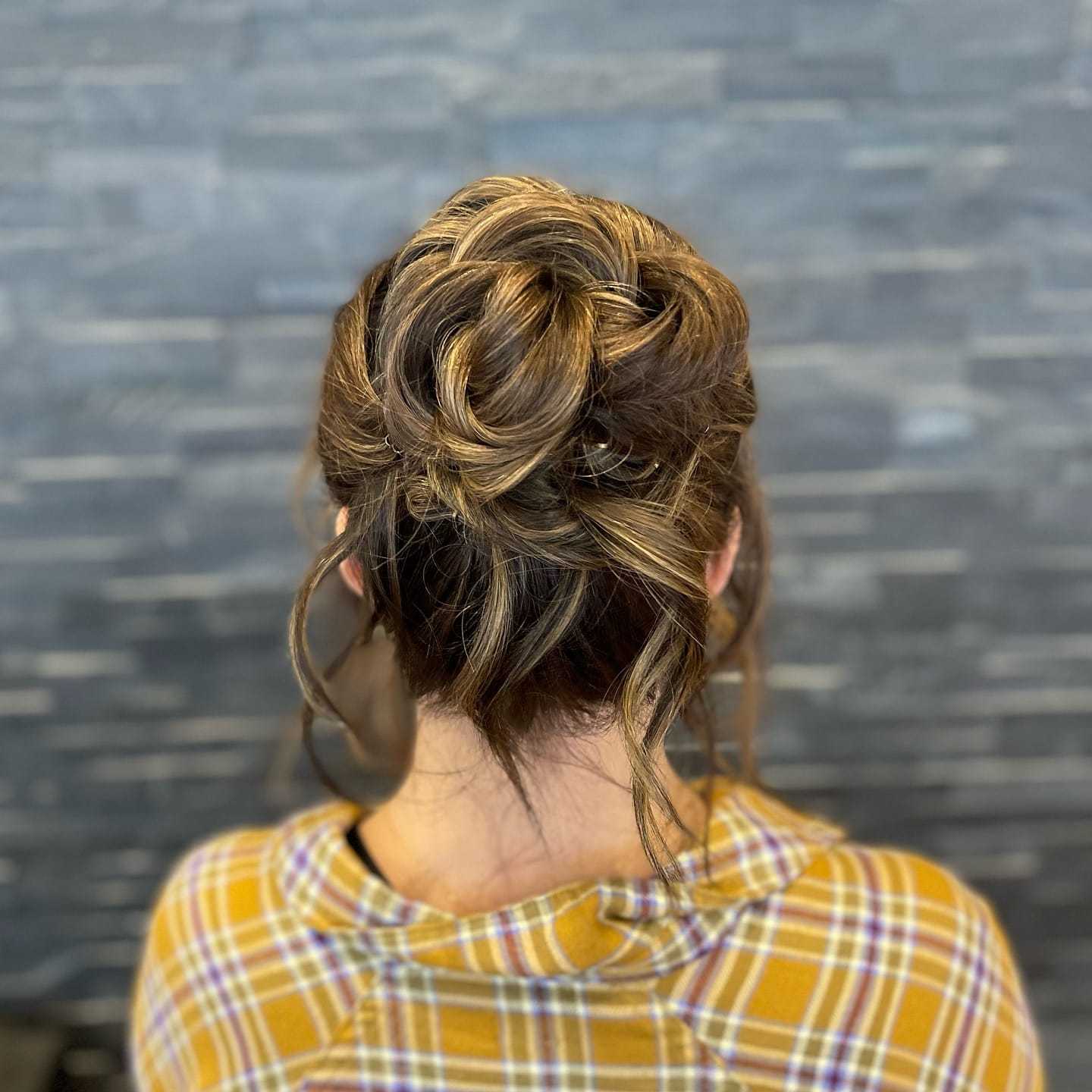 Woman with an intricately braided updo, wearing a yellow plaid shirt in front of a gray stone wall.