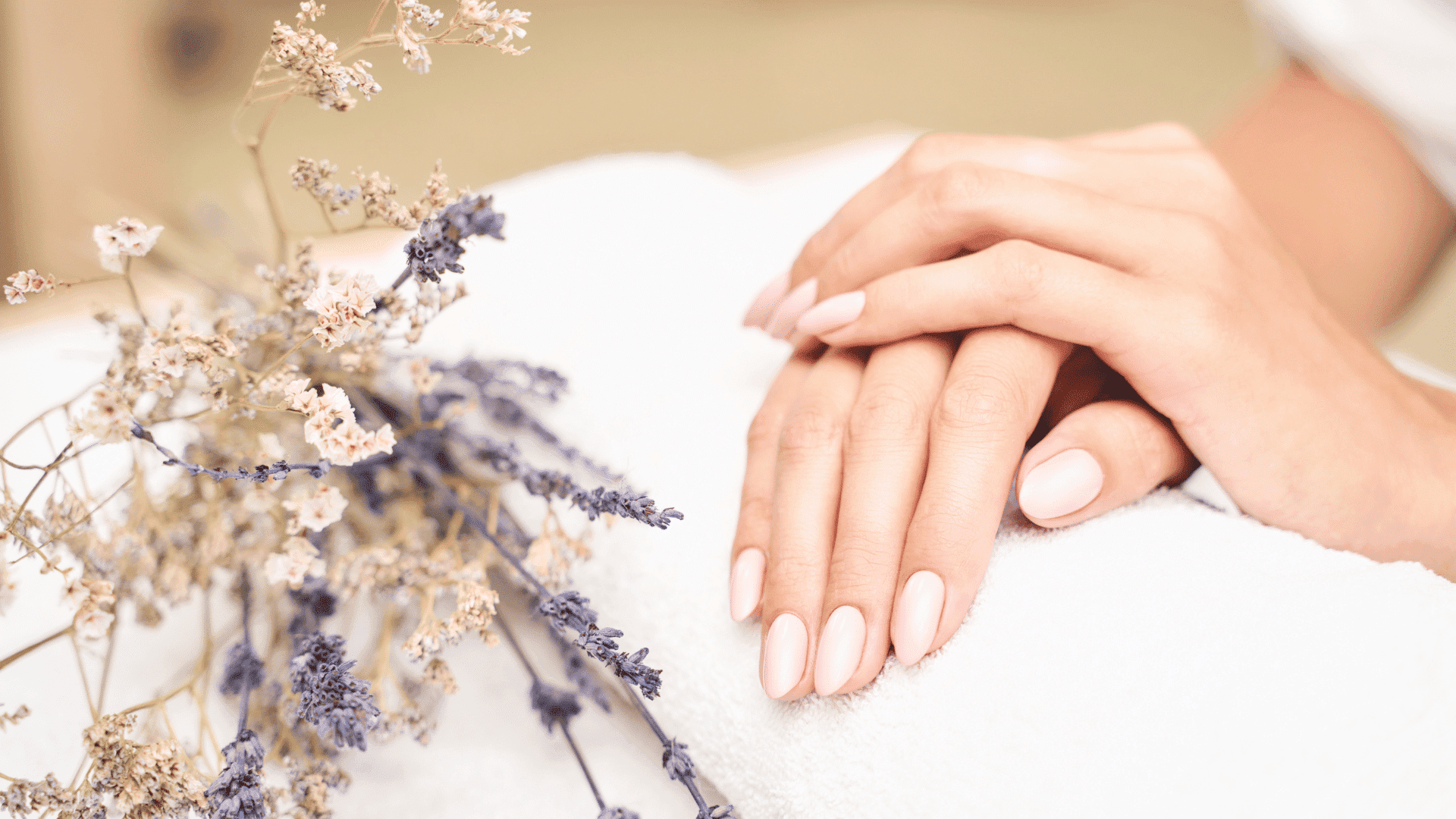 Hands with manicured nails resting on a towel beside dried lavender flowers.