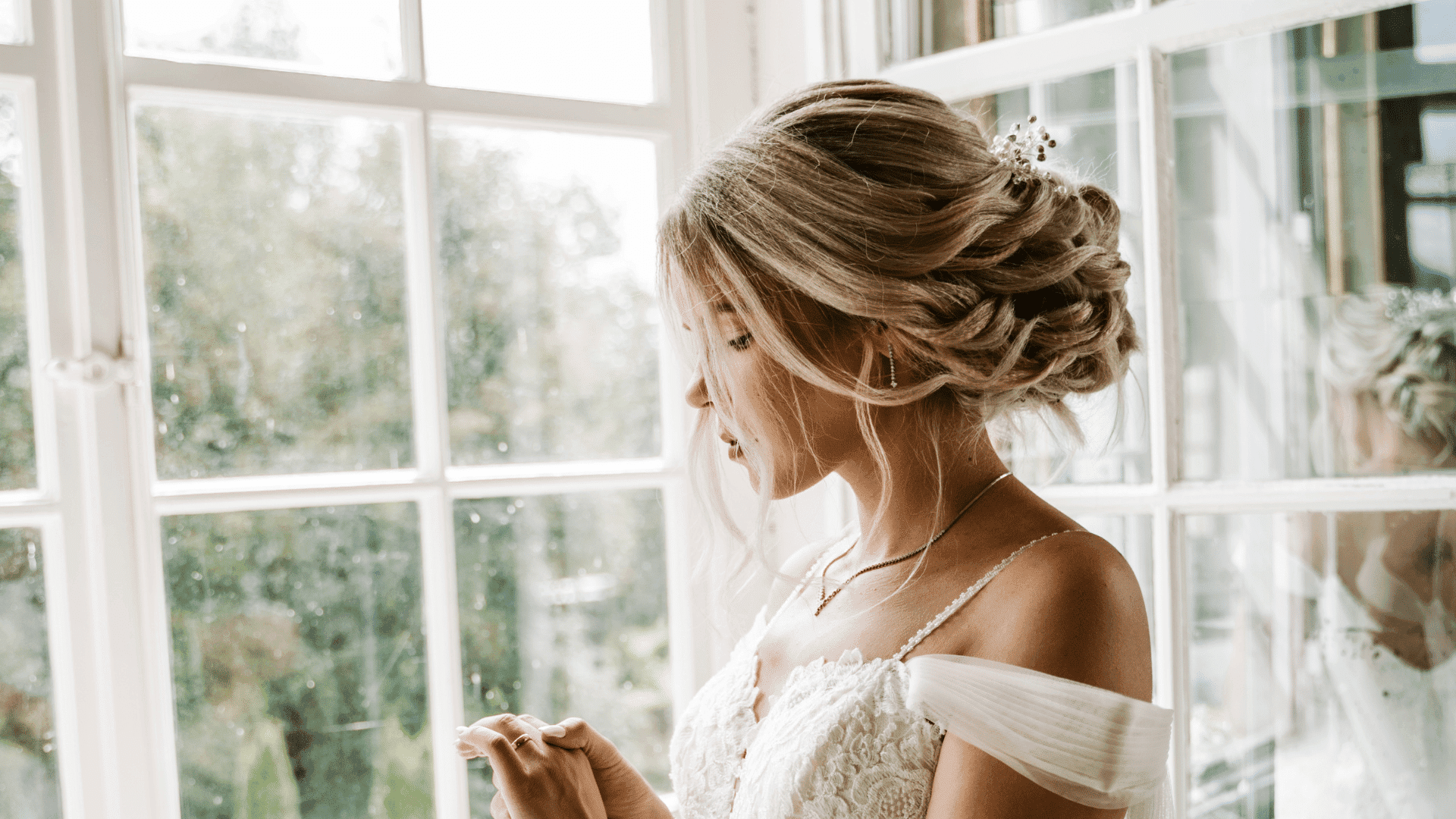 Bride in white dress and intricate updo holds a ring by a window, gazing thoughtfully at it.