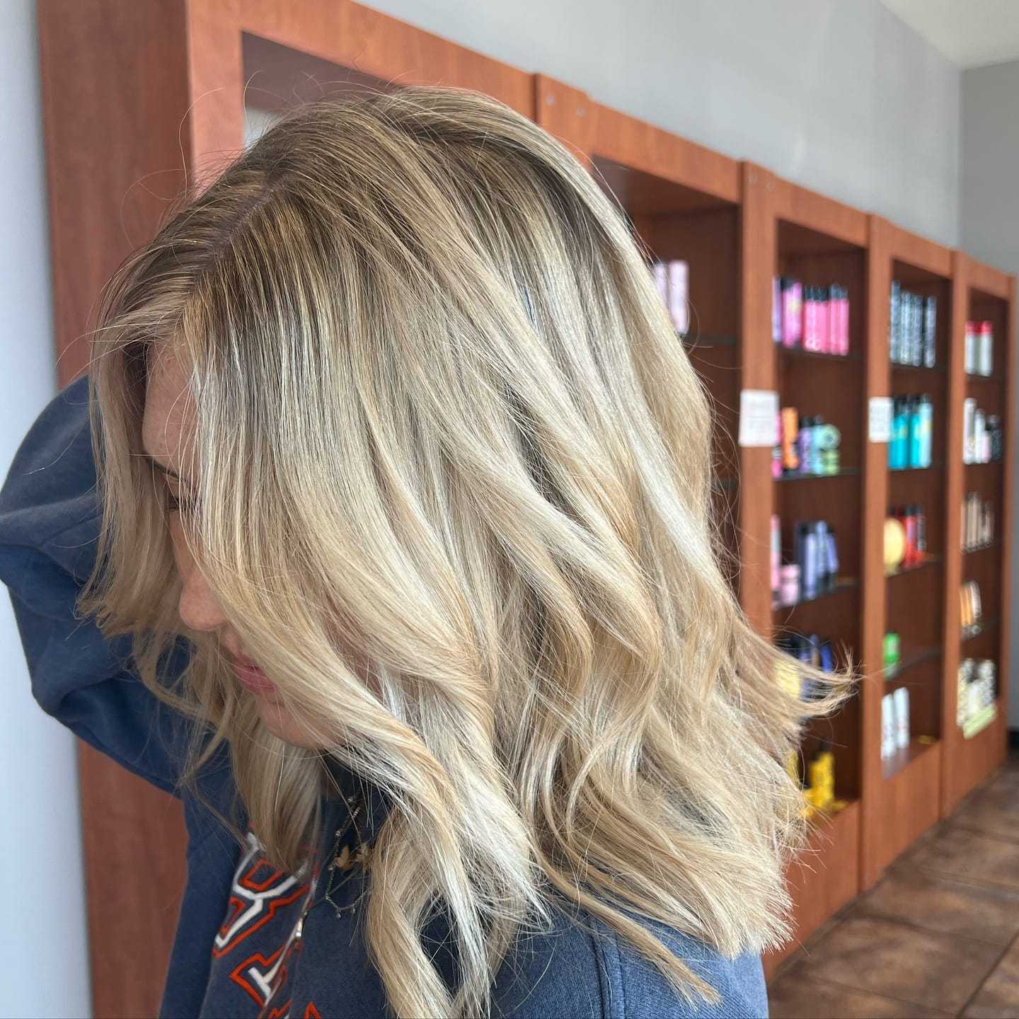 Woman with wavy blonde hair stands in front of salon shelves filled with hair products.
