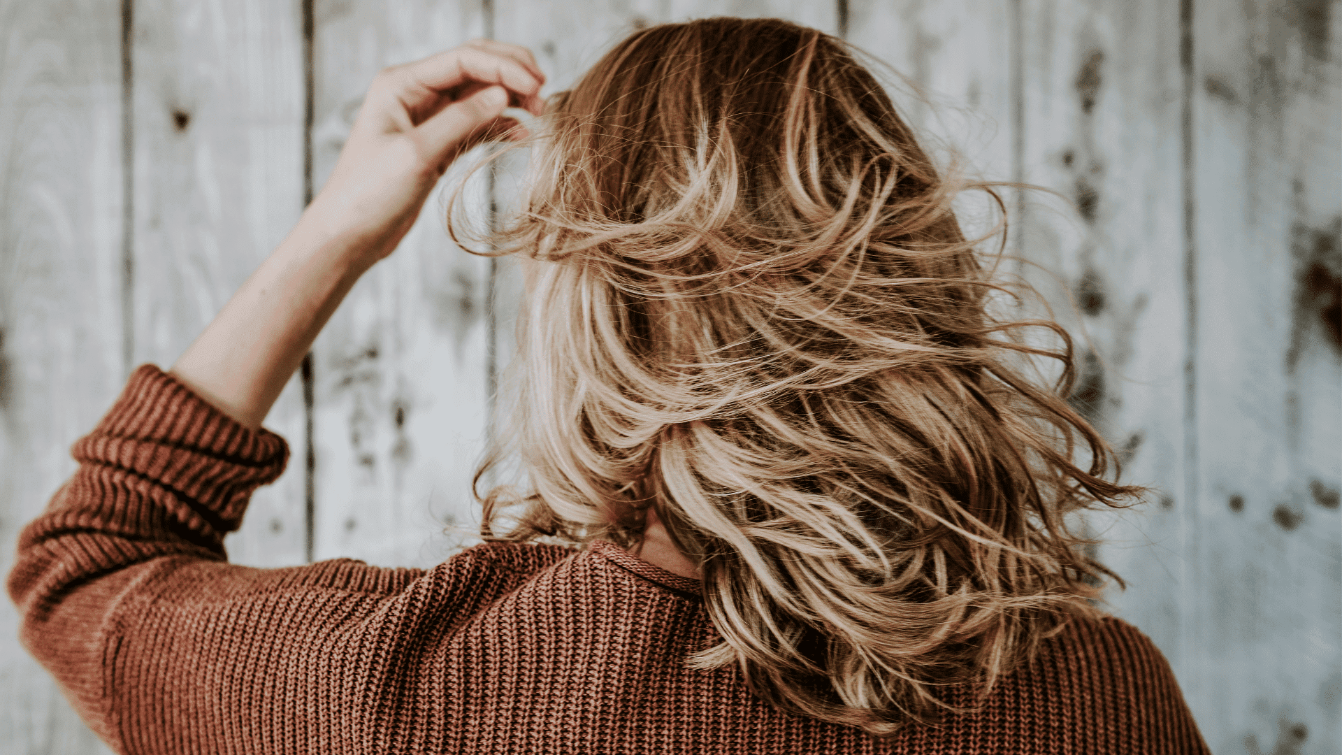 Woman with wavy blonde hair in a cozy sweater, touching her hair against a rustic wooden backdrop.